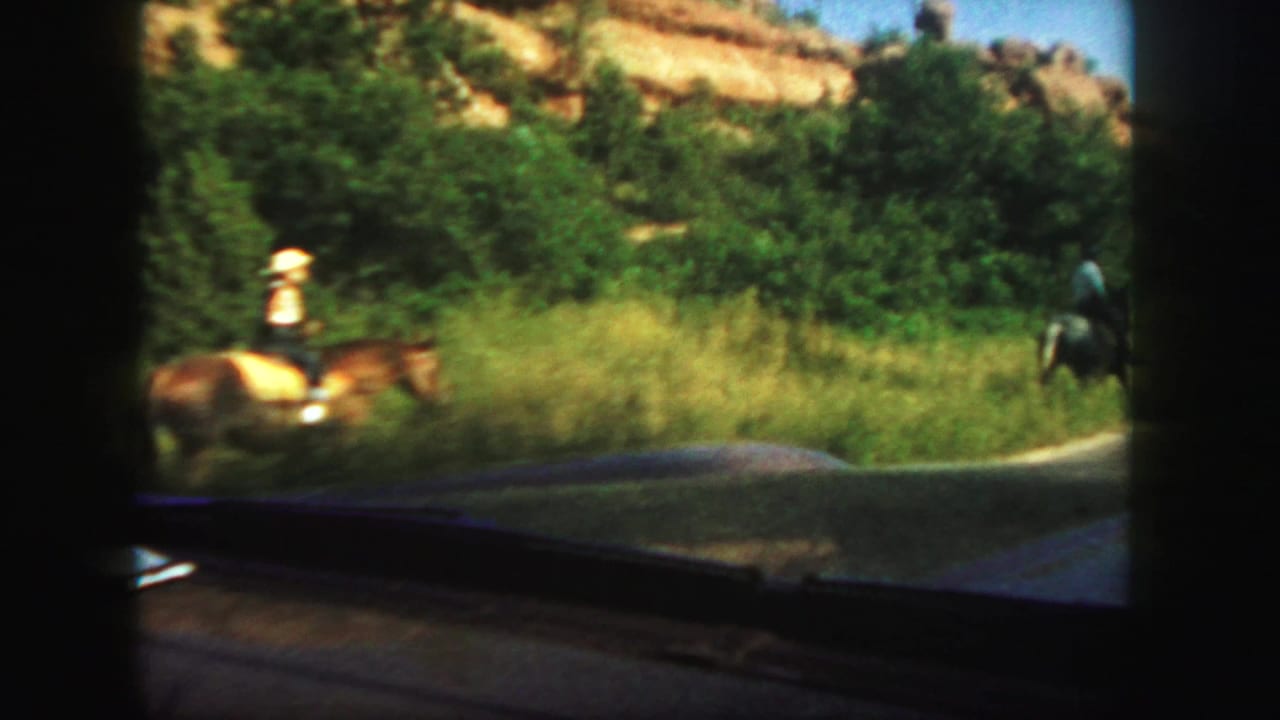 1958 Colorado home-movie view from inside a car as horseback riders move along a grassy roadside below rocky hills.