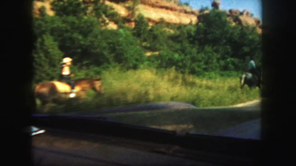 1958 Colorado home-movie view from inside a car as horseback riders move along a grassy roadside below rocky hills.
