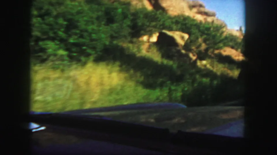 The moving car passes grassy brush and rock outcrops in Colorado, with the dashboard and windshield framing the 8mm view.