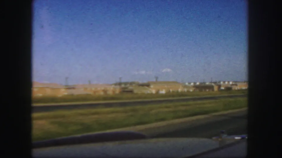 From inside the moving car, the view shifts to an open Colorado roadside with a paved highway, low hills, and distant buildings.