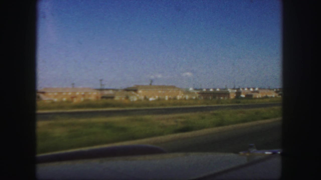 Distant factory buildings beyond a road and grass, seen from inside a moving car.