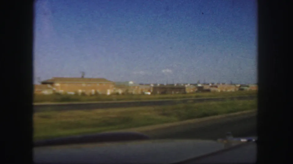 1958 Colorado drive-by view with low industrial structures, open grass, paved road, and car interior framing at the edge.