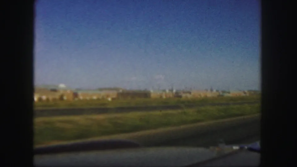 1958 Colorado 8mm frame with motion-blurred roadside grass, distant buildings, a paved road, and blue sky seen from a moving car.