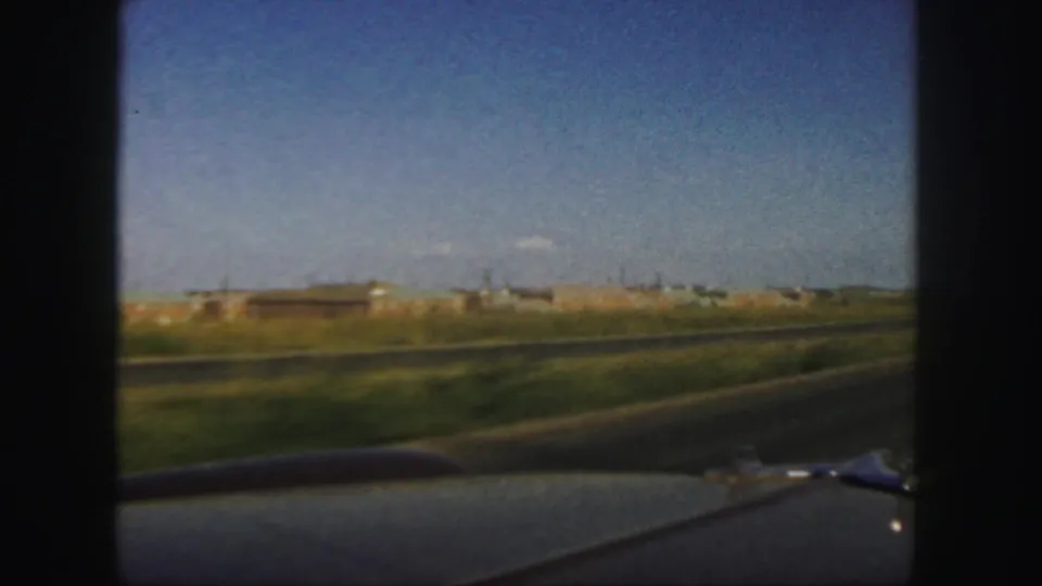 1958 Colorado home-movie frame showing a car-edge view of grassland, a road, and distant industrial buildings under blue sky.