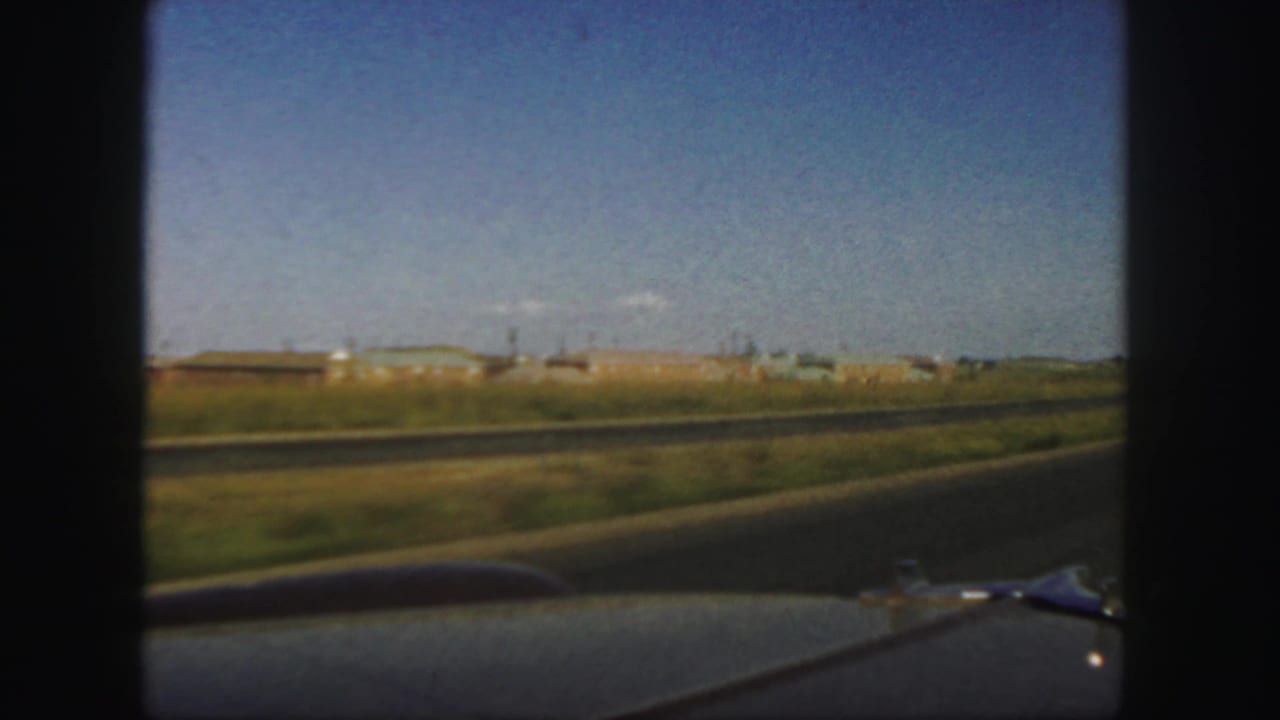 1958 Colorado road-trip frame from inside or just behind a moving car, looking past the hood toward a highway and low roadside buildings.