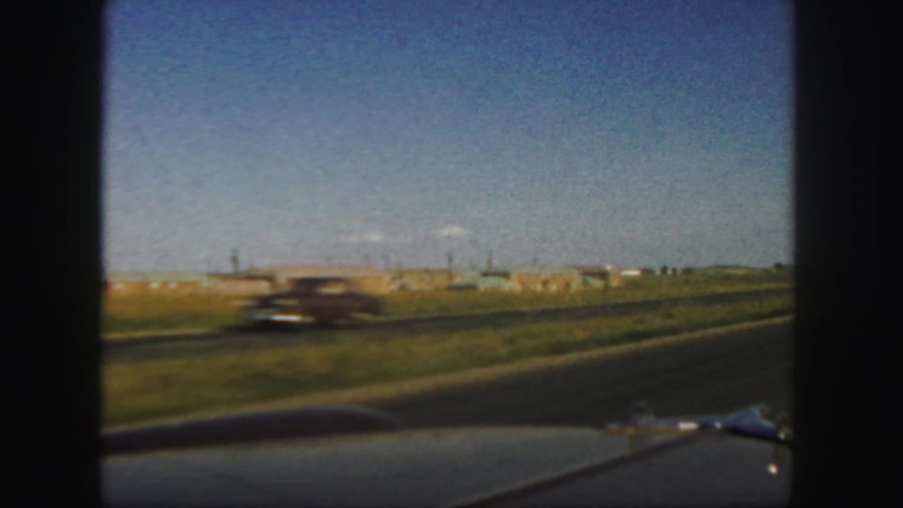 Roadside view with a blurred passing car, grassy verge, distant buildings, and blue sky.