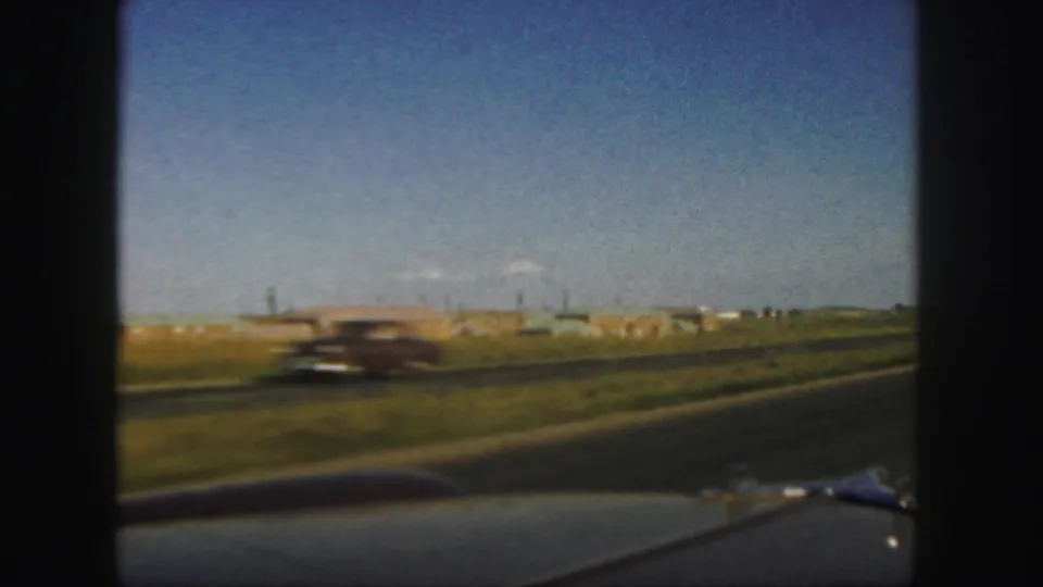 1958 Colorado highway view from a moving vehicle, with a blurred car passing across a grassy roadside and low buildings in the distance.