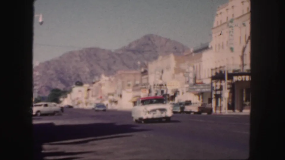 1958 Colorado small-town street scene with classic cars moving past storefronts and a visible hotel sign beneath a rugged mountain backdrop.