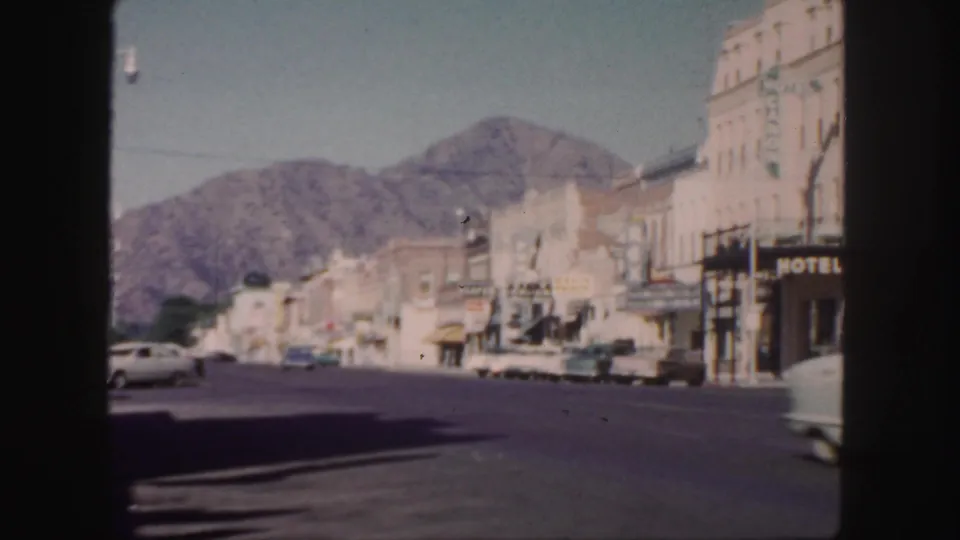 1958 Colorado main-street frame showing storefronts, a hotel sign, period cars, and mountains rising behind the commercial block.