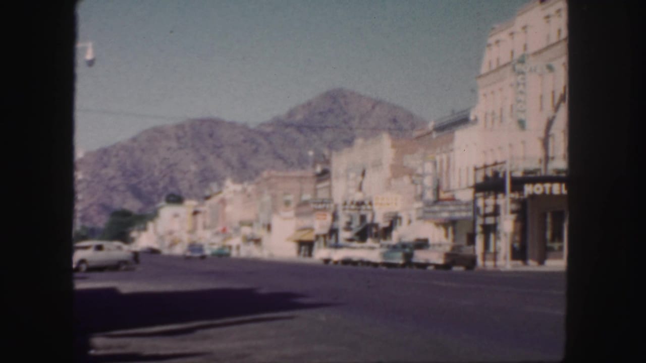 1958 Colorado street view from a vehicle or window, with mid-century cars, commercial buildings, a hotel sign, and mountains beyond town.