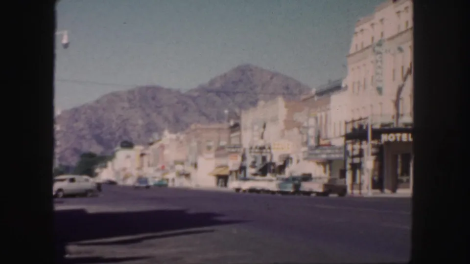 1958 Colorado street view from a vehicle or window, with mid-century cars, commercial buildings, a hotel sign, and mountains beyond town.
