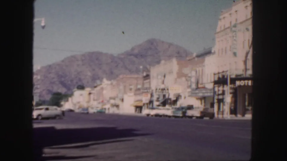 Colorado town street in 1958, seen through a dark foreground edge, with storefronts, parked mid-century cars, hotel signage, and a mountain backdrop.