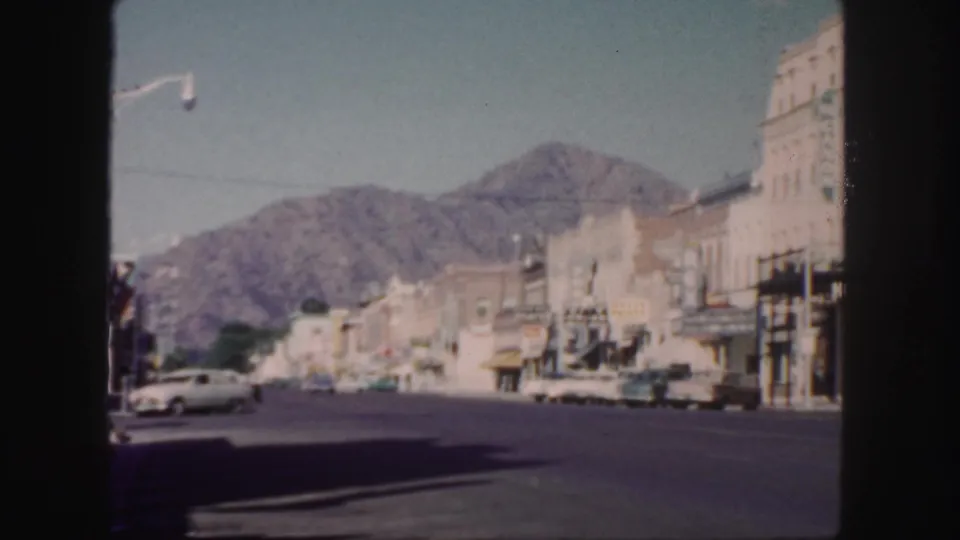 Late-1950s Colorado street scene with low commercial buildings, classic cars, a streetlight, clear sky, and mountains rising behind the town.