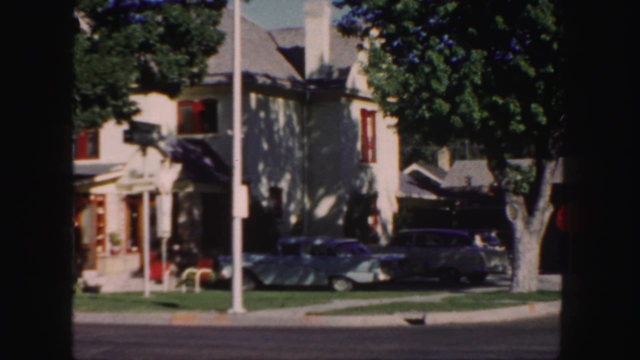 1958 Colorado home-movie frame showing a pale house with red trim, shade trees, two parked cars, lawn, curb, and street pole.