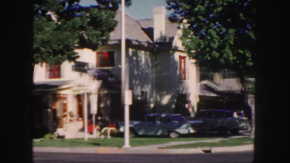 1958 Colorado exterior view of a light-colored residential house, red-trimmed windows, trees, parked cars, and a curbside street.