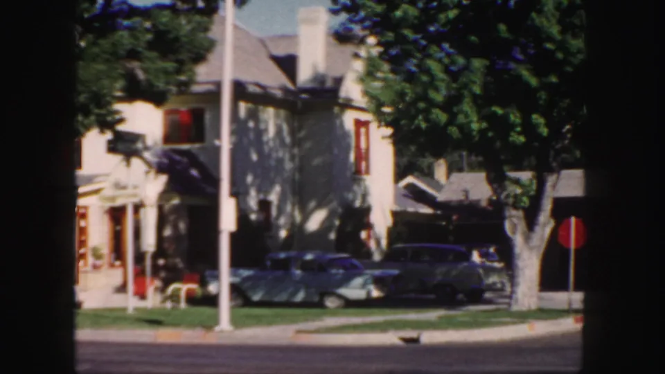 1958 Colorado home-movie frame with a pale house, large trees, parked cars, curb, and a red circular street sign near the driveway.