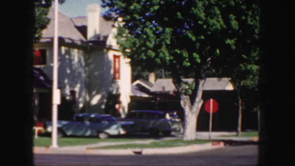 1958 Colorado street view showing a light house with red trim, leafy trees, two parked cars, a red sign, lawn, and dark 8mm frame edges.