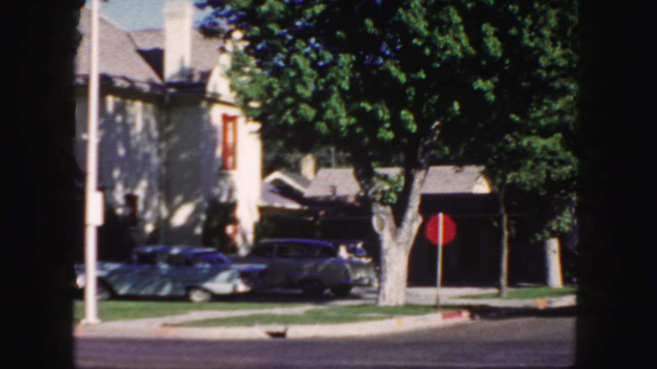 1958 Colorado 8mm frame showing a shaded residential corner with parked mid-century cars, a red stop sign, lawns, and houses.