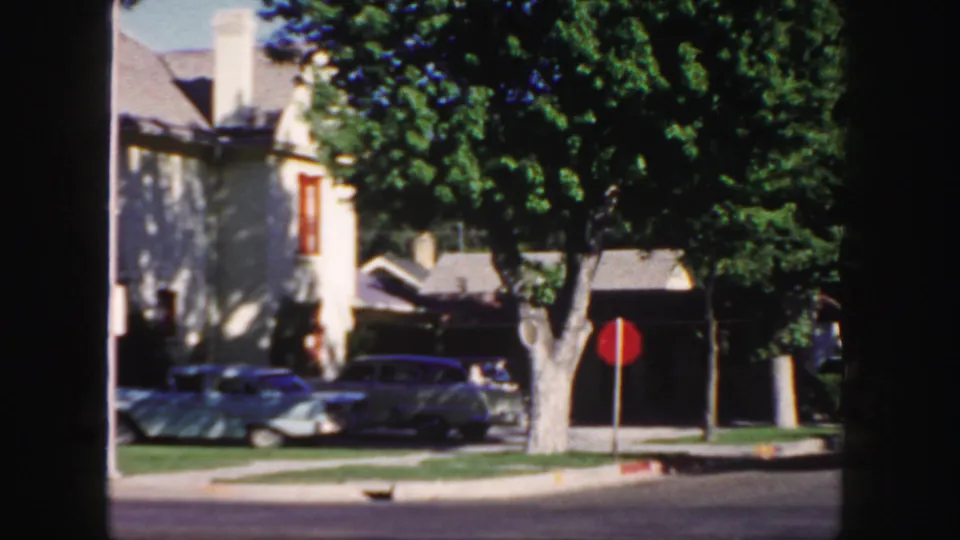 1958 Colorado residential street view with classic cars parked near a stop sign and houses partly shaded by trees.