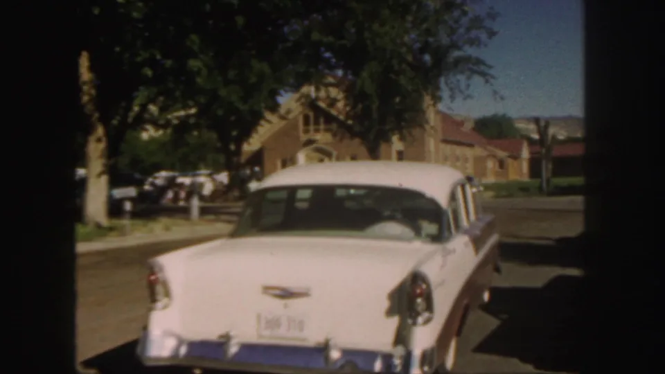 1958 Colorado 8mm frame from a moving car behind a white sedan, with residential buildings, trees, and parked vehicles ahead.
