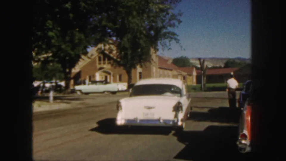 1958 Colorado street scene with a white sedan ahead, parked cars at the curb, houses, trees, and a distant person near the right edge.