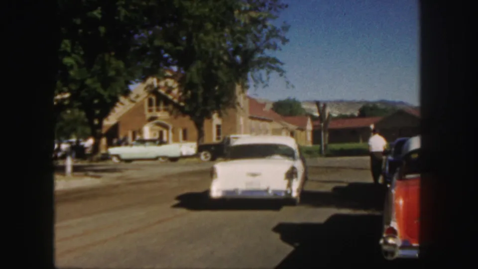1958 Colorado 8mm frame showing a white sedan on a road beside a brick church, with parked cars, trees, and distant mountains.