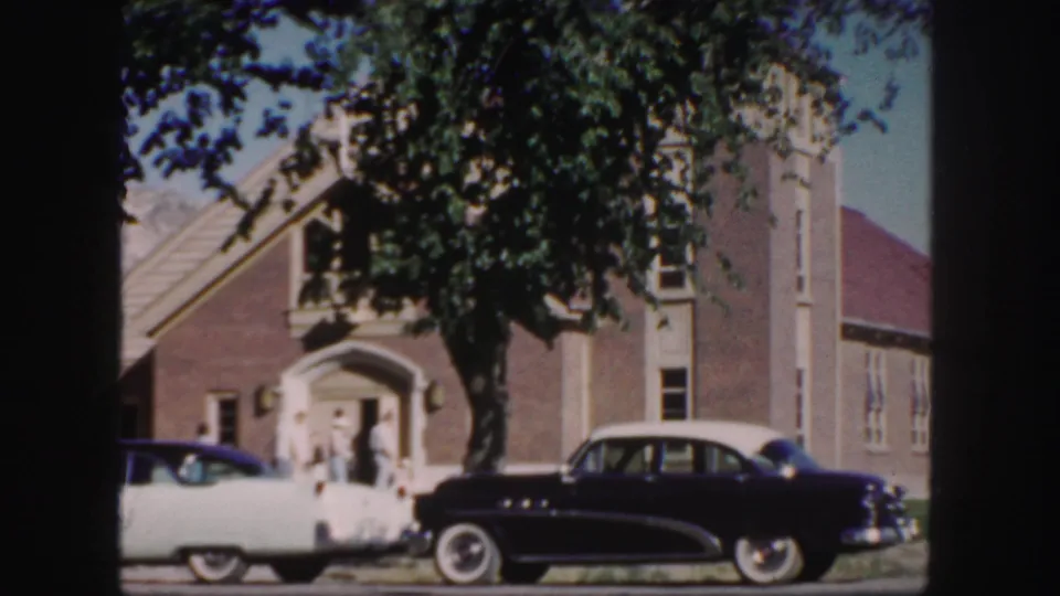 1958 Colorado frame showing a dark mid-century sedan and a light car parked outside a brick church with people near the entrance.