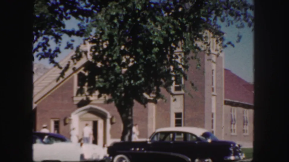 1958 Colorado church exterior with a black sedan, a white car, tree shade, and small figures near the doorway.