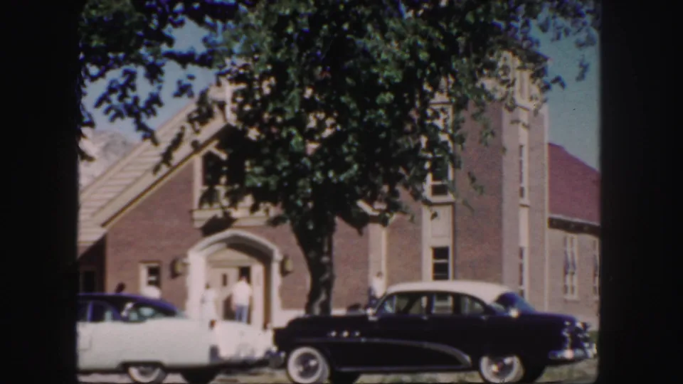 Home-movie frame of a brick church in Colorado, with mid-century cars at the curb and people by the entrance.