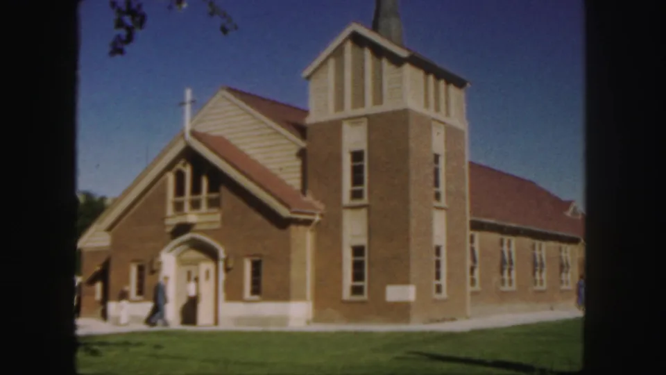 1958 Colorado 8mm frame showing a brick church with red-tiled roof, steeple, cross, lawn, and people standing near the entrance.