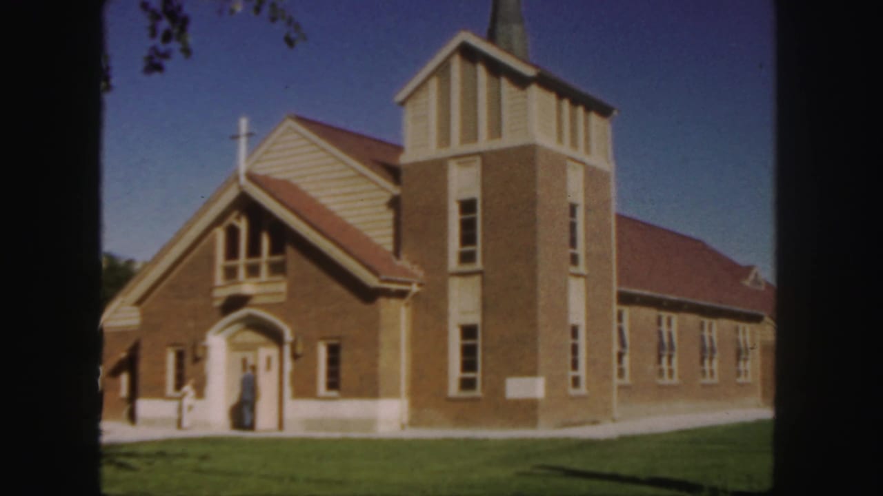 Church facade with steeple, cross, red roof, side windows, lawn, and people by the entrance.