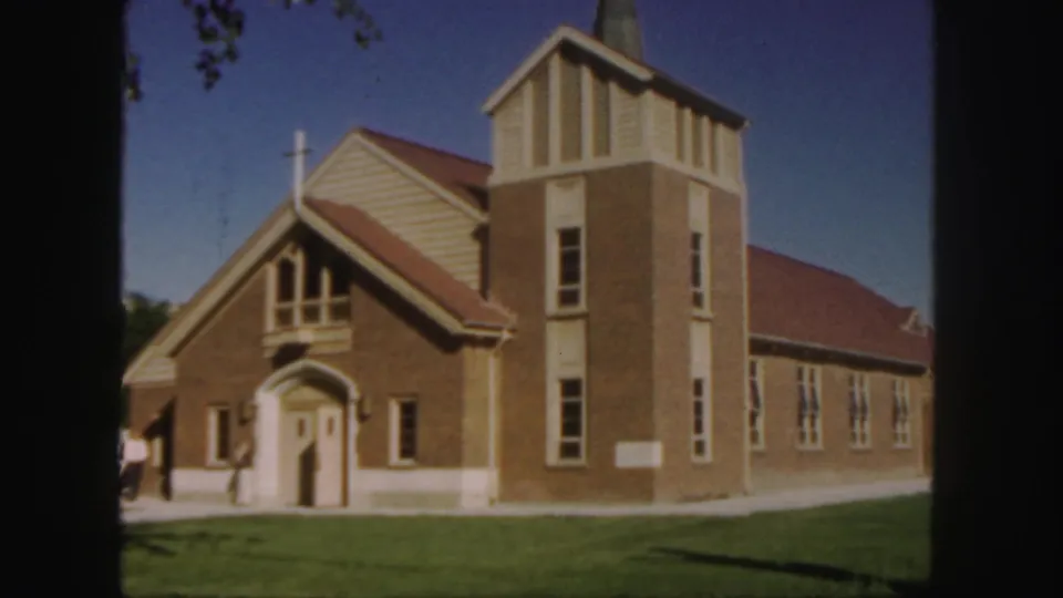 1958 Colorado frame showing the brick church from an exterior angle, with steeple, cross, red roof, arched entrance, and people nearby.
