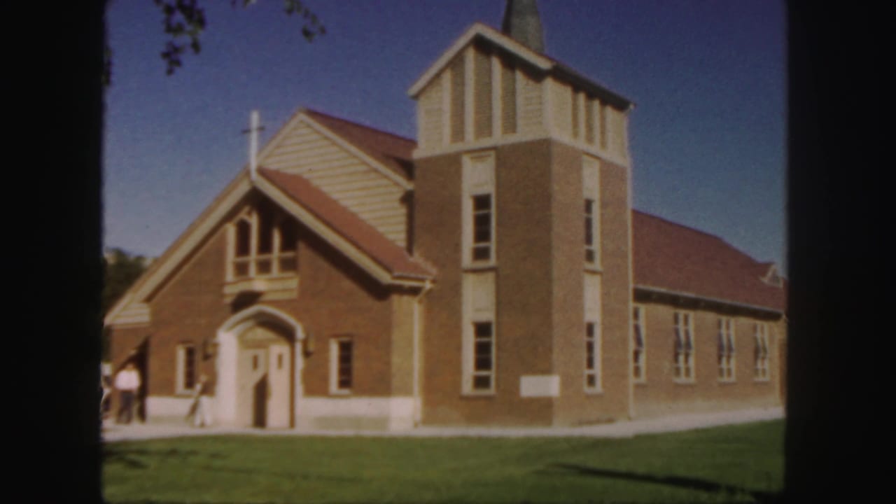 Brick church with steeple, cross, lawn, and people near the entrance.