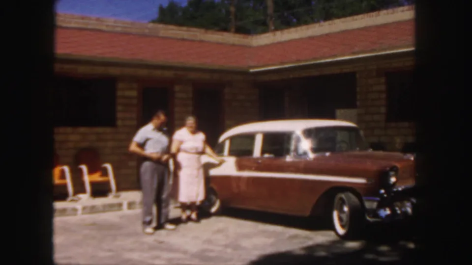 1958 Colorado home-movie frame showing a man and woman posed beside a brown-and-white classic car outside a brick building.