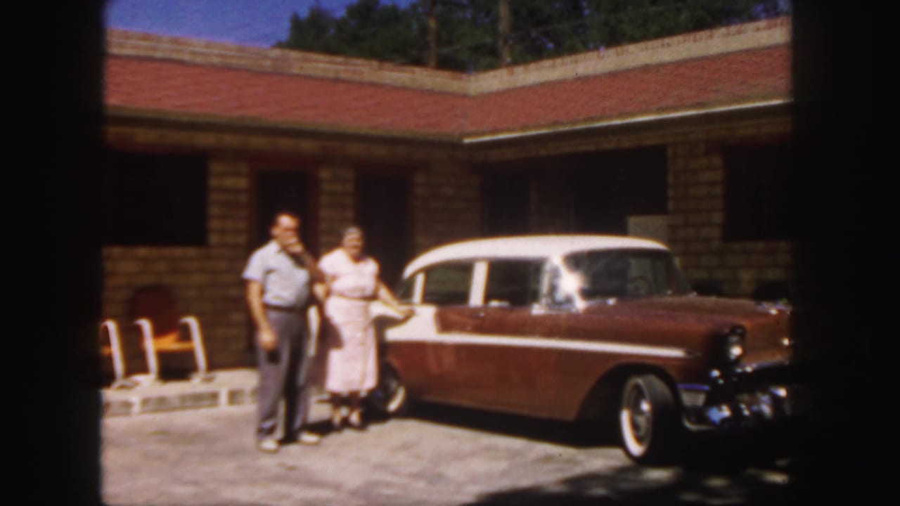 In 1958 Colorado home-movie footage, a couple stands by a two-tone station wagon in bright sun outside a brick motel.