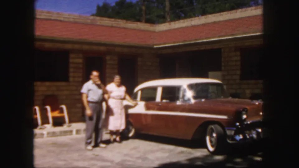 In 1958 Colorado home-movie footage, a couple stands by a two-tone station wagon in bright sun outside a brick motel.