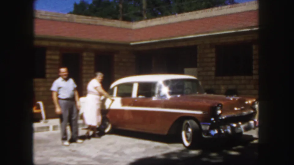 A 1958 Colorado home-movie frame shows the couple beside the parked station wagon, with the motel facade behind them.