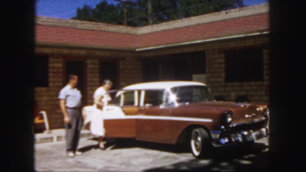 In 1958 Colorado home-movie footage, two adults stand beside a brown-and-white sedan with an open door near a brick building.