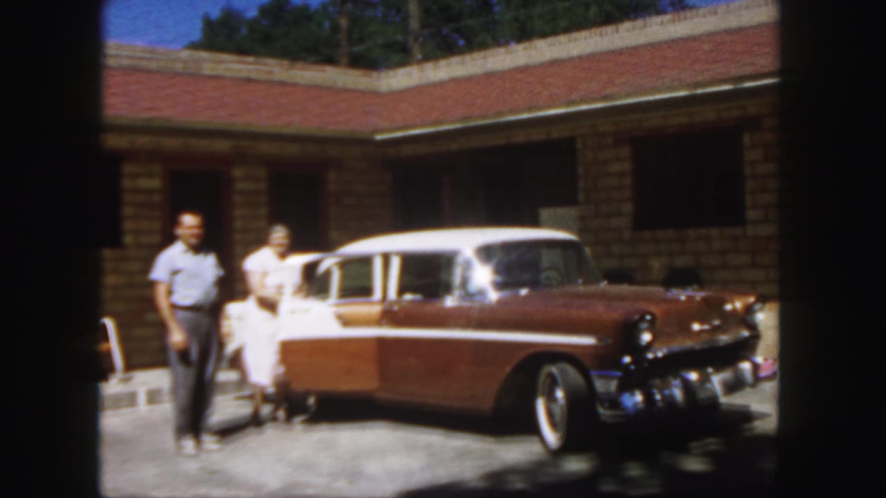 Man and woman stand near the open side of a two-tone sedan in front of a brick building.