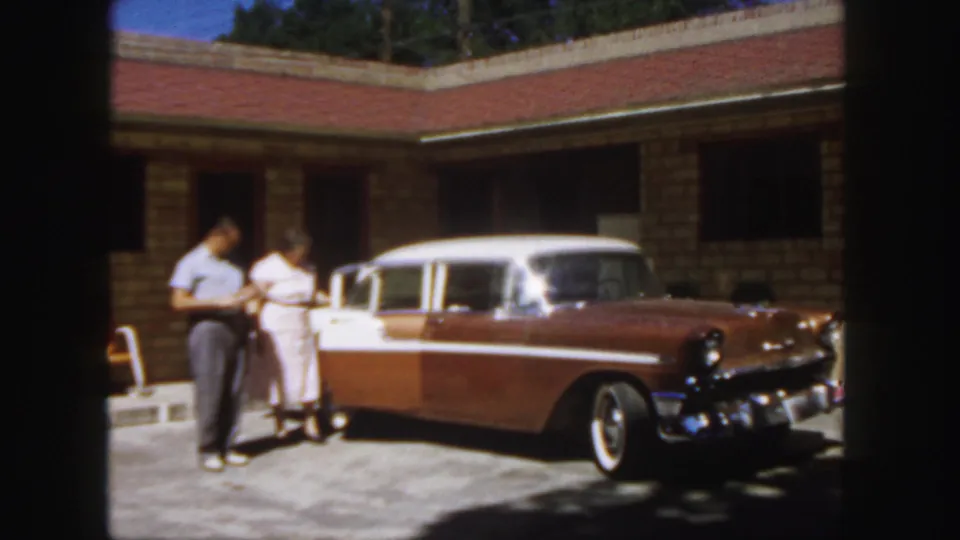 The man and woman appear to look toward the car door area beside the 1950s sedan in the Colorado home-movie scene.