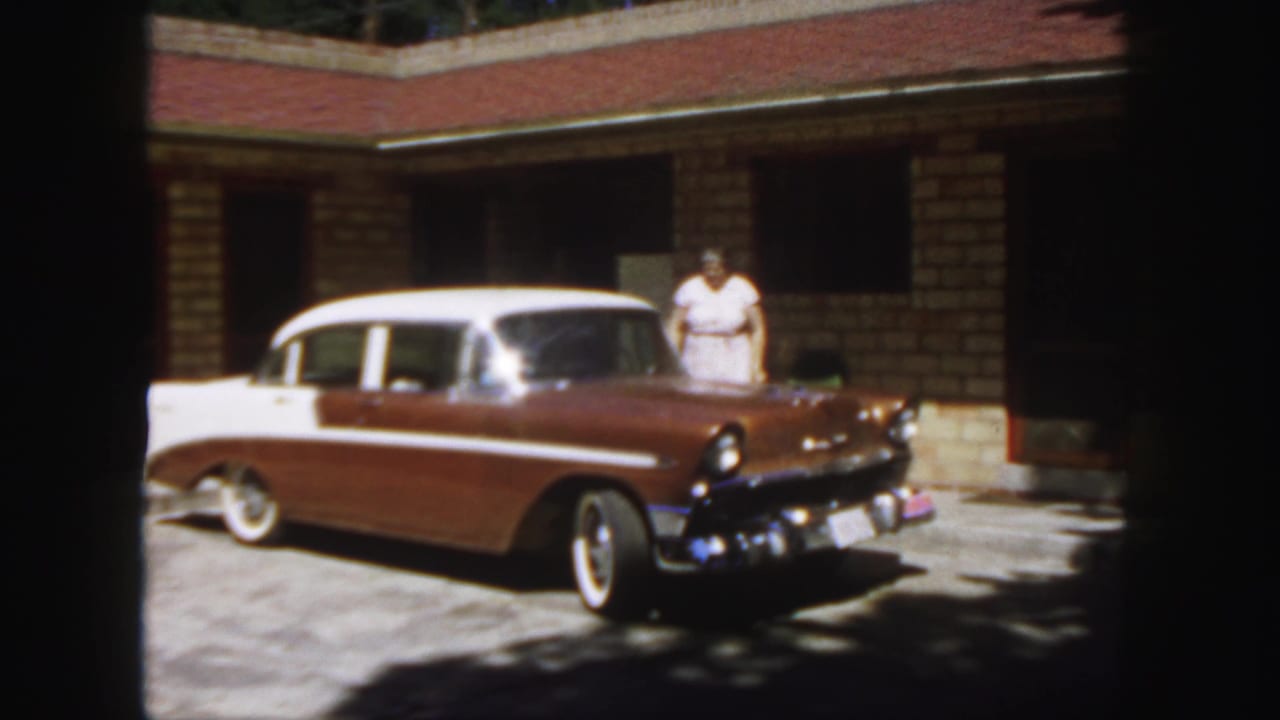 Brown and white mid-century sedan parked near a brick building with an adult in a white dress nearby.