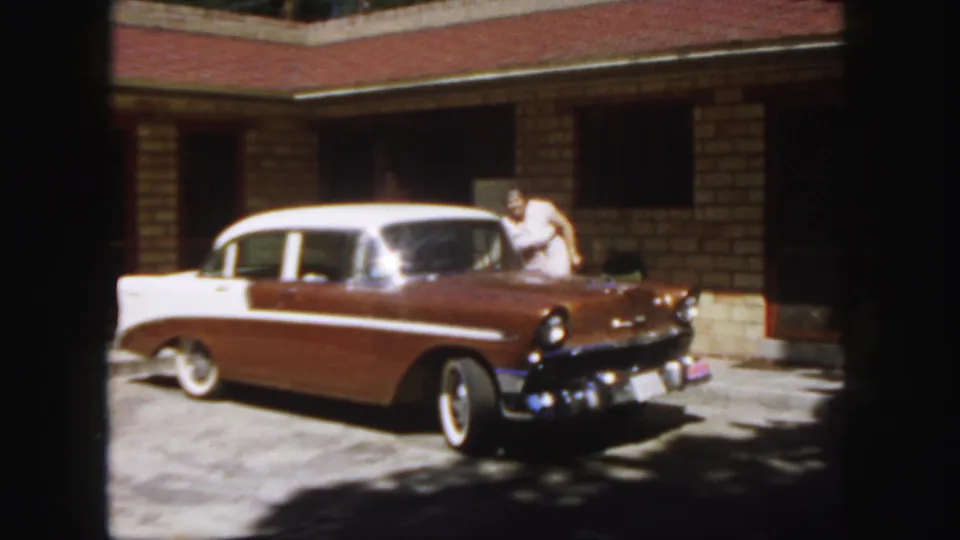 1958 Colorado home-movie frame with the brown and white sedan in front of a brick building and an adult close to the passenger-side window.
