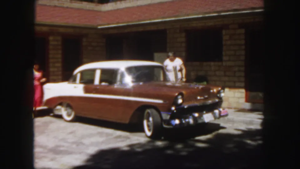 1958 Colorado 8mm frame with a two-tone sedan, an adult in white by the front passenger area, and another adult in red partly visible behind the car.