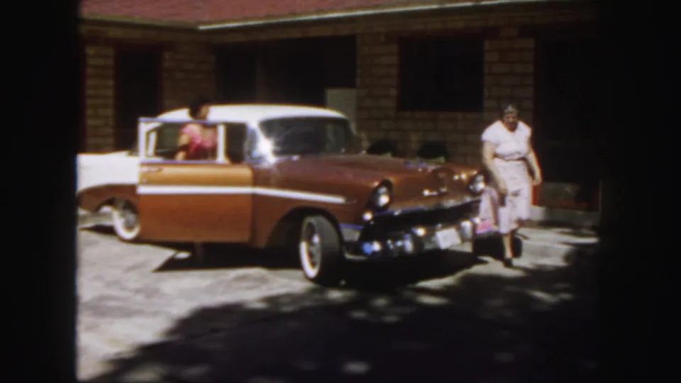 1958 Colorado frame showing one woman inside the sedan and another walking near the front in a sunny driveway.
