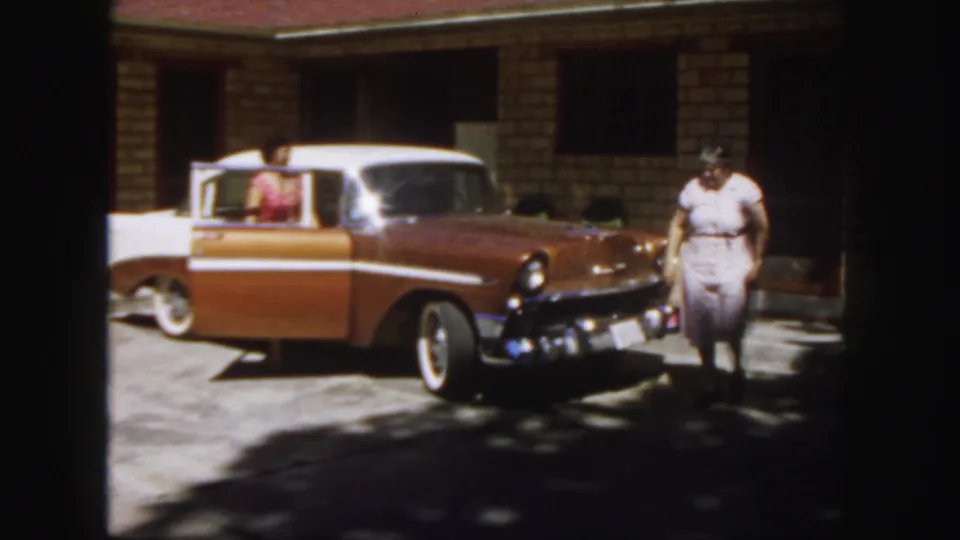 1958 Colorado home-movie frame showing a woman near a two-tone sedan with an open rear door while a passenger sits inside.