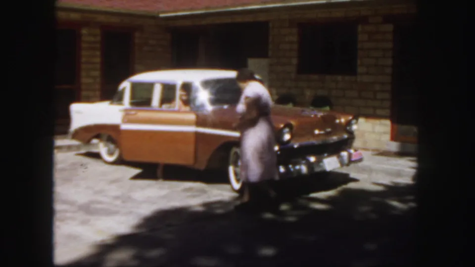 1958 Colorado frame with a woman moving along the front of a two-tone sedan, with another person visible through the open rear door.