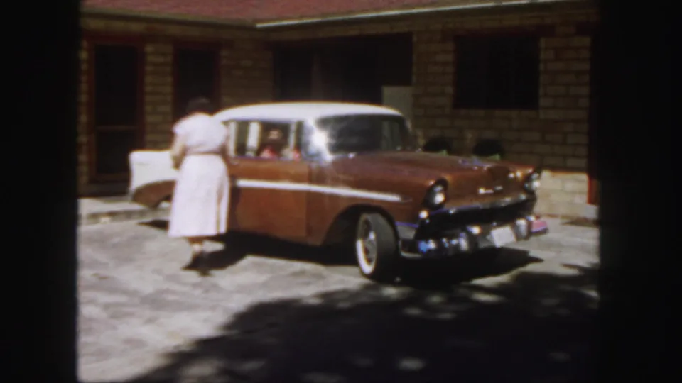 1958 Colorado home-movie frame showing a woman at the side of a parked two-tone sedan with its rear door open.