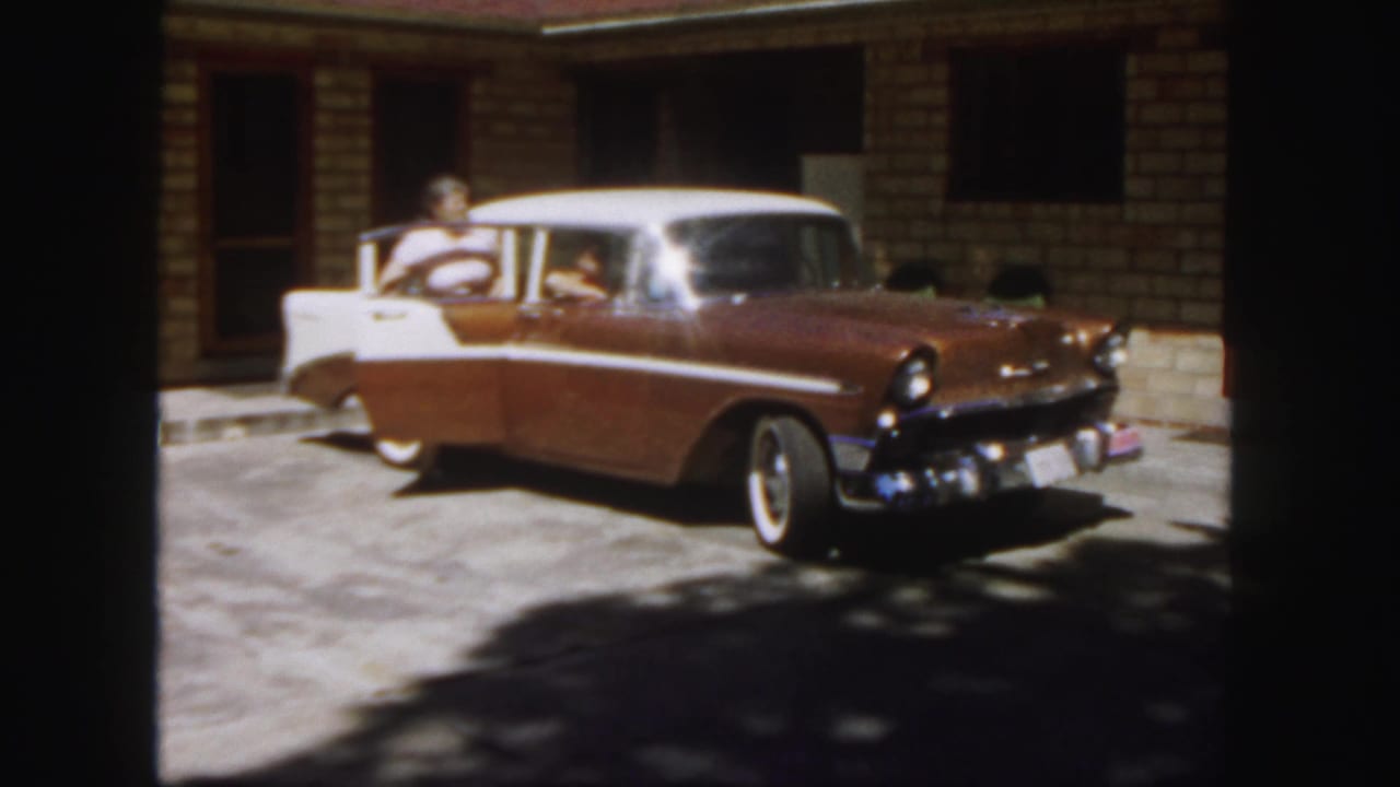 In 1958 Colorado footage, a man in a light shirt stands by a two-tone Chevrolet near a building entrance.
