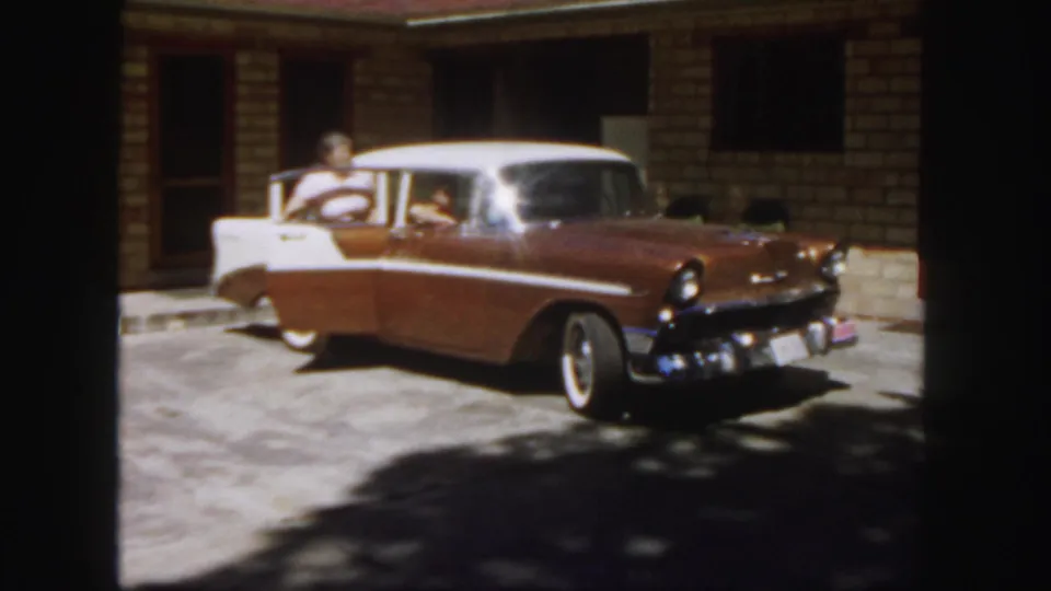 In 1958 Colorado footage, a man in a light shirt stands by a two-tone Chevrolet near a building entrance.
