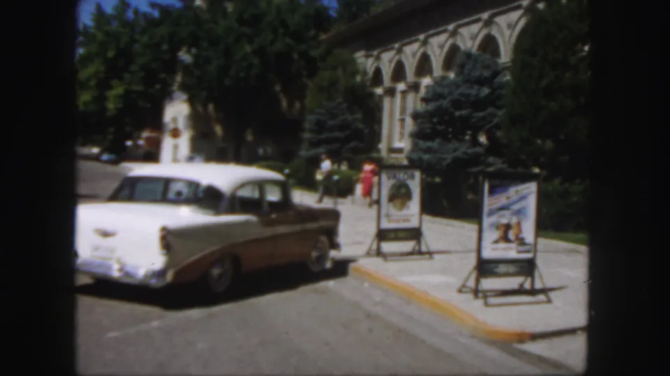 The car is seen from behind as it passes poster boards and pedestrians near a midcentury Colorado building.